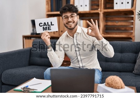 Hispanic man with beard working at therapy office holding call me sign doing ok sign with fingers, smiling friendly gesturing excellent symbol 