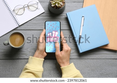 Bonus gaining. Woman using smartphone at grey wooden table, top view. Illustration of gift boxes, word and falling confetti on device screen