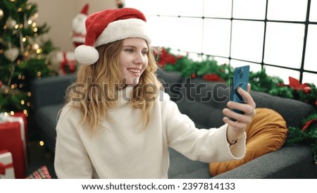 Young blonde woman making selfie by smartphone sitting on sofa by christmas tree at home