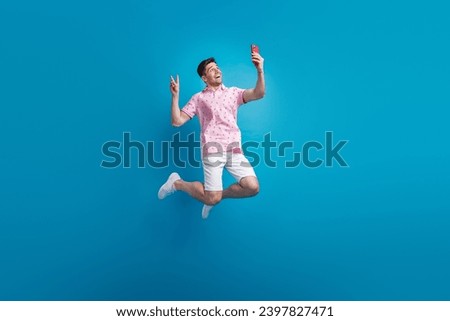 Full size photo of excited handsome guy jump hold smart phone make selfie show v-sign isolated on blue color background