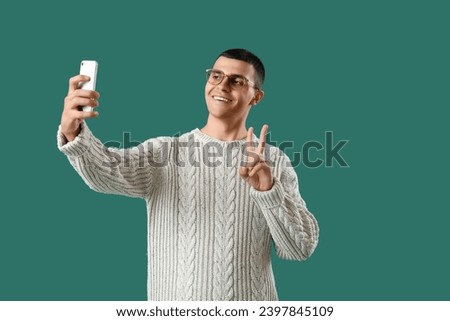 Young man in knitted sweater taking selfie on green background