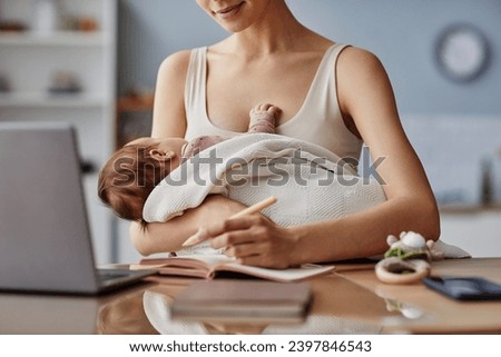 Cropped shot of mother writing down ideas in front of computer and holding her baby in white blanket, copy space