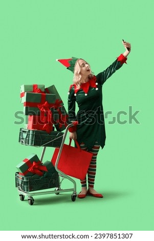 Happy young woman dressed as elf with shopping cart and Christmas gifts taking selfie on green background