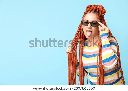 Portrait of fashionable young woman with dreadlocks showing tongue on blue background Royalty-Free Stock Photo #2397863569