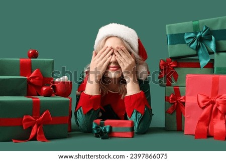 Emotional young woman dressed as elf with Christmas gifts on green background
