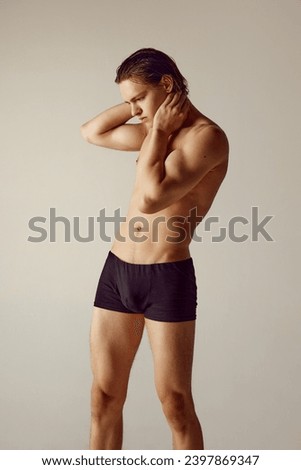 Handsome, shirtless, muscular young man fit fit body standing in underwear, holding hand on neck against grey studio background. Concept of men's beauty, health, body care, sportive lifestyle Royalty-Free Stock Photo #2397869347