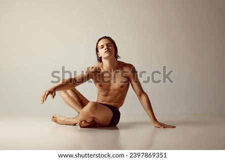 Handsome young guy with sportive, muscular body sitting on floor, posing shirtless in underwear against grey studio background. Concept of men's beauty, health, body care, sportive lifestyle Royalty-Free Stock Photo #2397869351
