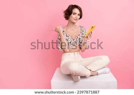 Full size photo of optimistic girl dressed blouse sit on cube hold smartphone directing empty space isolated on pink color background