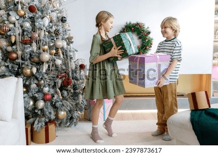 girl and boy have fun near Christmas tree, sister loads her brother by pile of colorful presents.