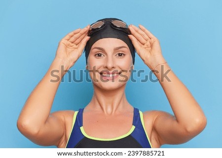 Sportsgirl with bathing cap holding hands on her goggles, stands isolated on blue background, sport concept, copy space. 