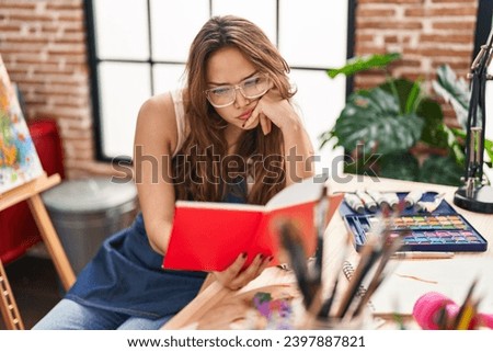 Young beautiful hispanic woman artist reading book sitting on table at art studio