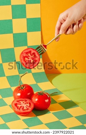 The hand is holding a metal fork pinned to half of a tomato. Juicy red tomatoes are displayed on a checkered background with two main colors: green and yellow. Art space with fresh vegetables.