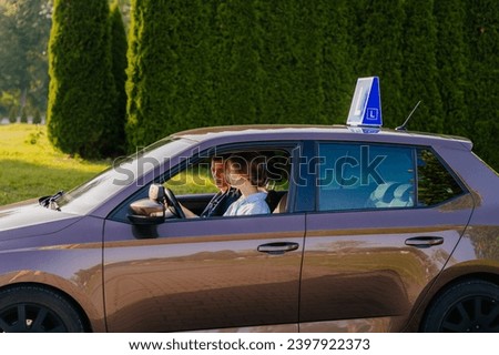 Carefully instructor sitting in a learning car next to a female student driver. Happy woman drives brown left-hand driving vehicle with blue L plate on a roof.