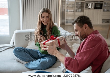 Pleasantly smiling merry woman looking with excitement as intrigued interested man trying to unwrap gift box with red ribbon. Unexpected surprise for birthday, anniversary, New Year, Christmas Royalty-Free Stock Photo #2397925781