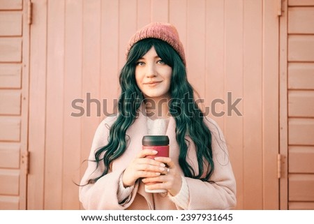 Stylish young smiling hipster woman with color hair wearing trendy peach color coat and hat with reusable coffee cup on wooden background. Urban seasonal city street fashion. Color of the 2024 year. Royalty-Free Stock Photo #2397931645