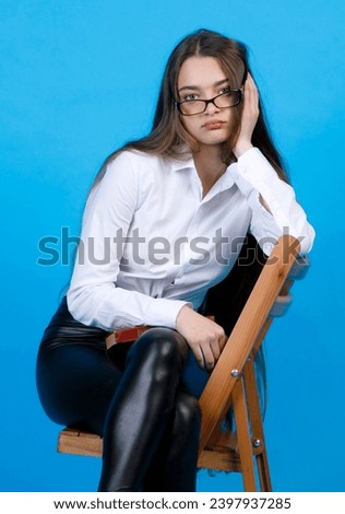 Bored caucasian woman in glasses, resting head on hand, while sitting on chair indoor. Portrait of upset girl in black leggings, sitting cross-legged, isolated on blue background. Concept of emotions.