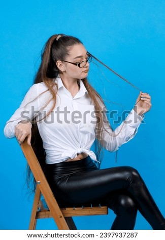Upset caucasian woman in glasses, touching long hair, while resting on chair. Portrait of bored cute girl in white shirt, sitting cross-legged, isolated on blue studio background. Concept of emotions.