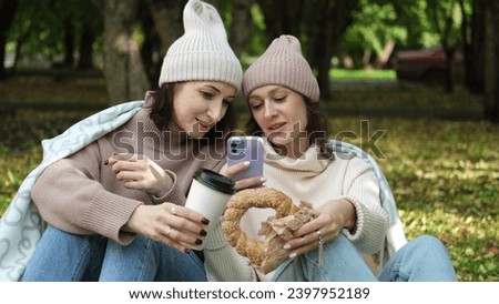 In autumn, two cheerful girls walk through the park during the day, drink coffee and take pictures of glasses of coffee and bagels against the background of autumn foliage for social networks.