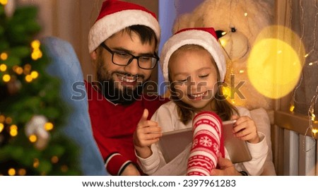 Happy family father and little daughter in red santa hats use tablet and laugh sitting by Christmas tree. New Year Christmas holiday time.