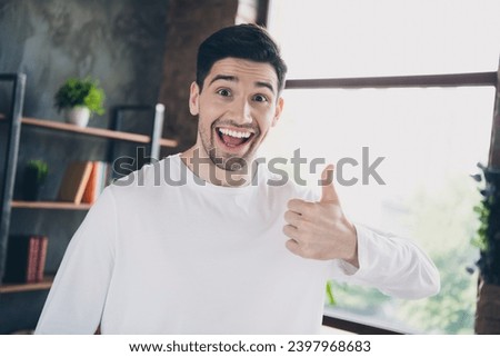 Photo of excited impressed young guy dressed white shirt open mouth showing thumb up indoors house room