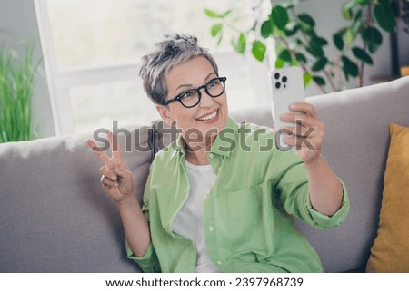 Photo of pensioner gray haired woman wearing green shirt making self portrait using smartphone show v sign isolated at home in living room