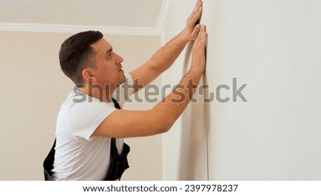 Professional Caucasian worker glues a strip of wallpaper on the wall. Man with dark hair in a uniform glueing wallpaper. Royalty-Free Stock Photo #2397978237