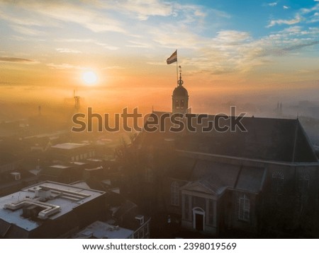 Drone view across town of Sneek with Dutch flag on top of church at sunrise. Bright orange colours and little fog or mist in the streets of the city. High quality photo