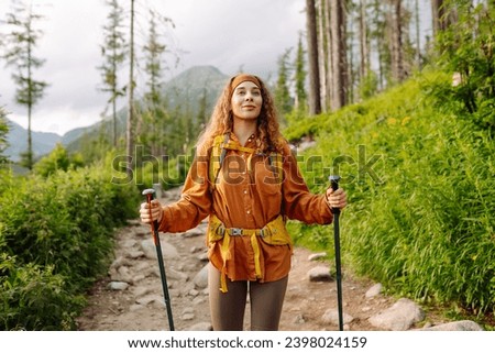 Young female traveler with hiking poles and a bright backpack enjoys the mountain scenery along a hiking trail. Active lifestyle. Wanderlust.