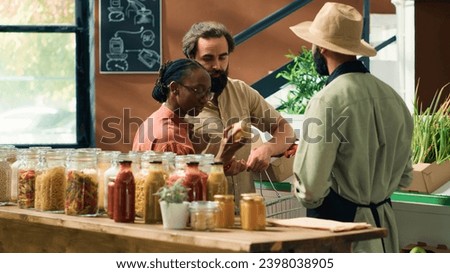 Merchant assists customers in grocery store, showing freshly harvested fruits and vegetables. Young couple searching for organic produce at local zero waste supermarket, sustainable lifestyle. Royalty-Free Stock Photo #2398038905
