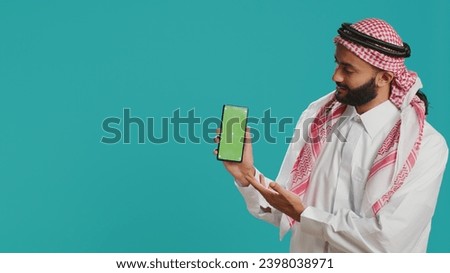 Middle eastern man points at greenscreen display on smartphone, showcasing isolated copyspace template on mobile device screen. Young person in islamic costume shows blank chromakey.
