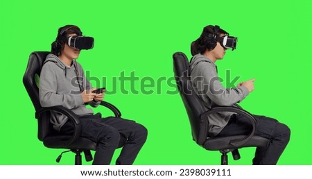 Young guy engages in web based gameplay clash with other people while playing video games using multiple devices against greenscreen background. With virtual reality glasses, man looks joyful.