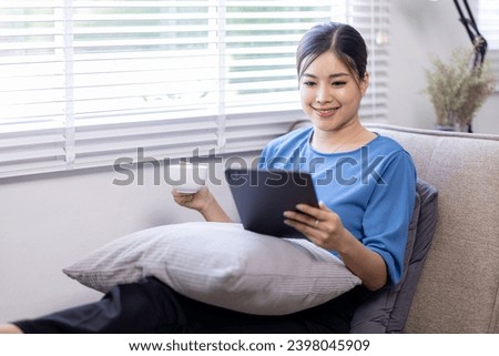 Asian young woman sitting on the yellow sofa using digital tablet and coffee cup and learning shopping or working online near window