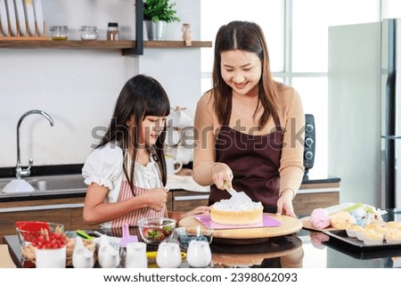 Asian beautiful female baker pastry chef mother wears apron standing smiling helping teaching little girl daughter decorating cake with whipping cream making homemade bakery in decorated home kitchen. Royalty-Free Stock Photo #2398062093