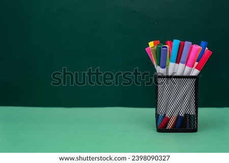 A bunch of colored pens in the square metal pen holder sitting on green surface