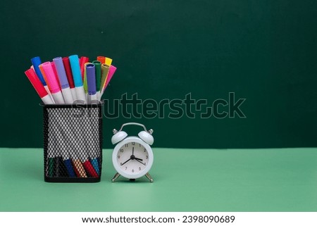 A bunch of colored pens in the square metal pen holder sitting next to white alarm clock on green surface