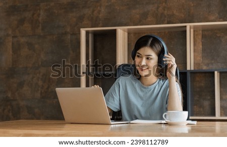 Young female student wearing headphones sits intently and happily studying online on her laptop in the living room at home. Royalty-Free Stock Photo #2398112789