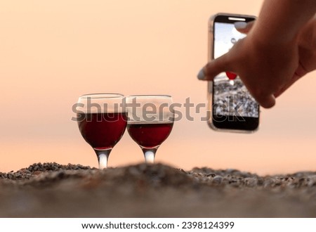 A girl takes a picture of a glass of wine on her phone at sunset on the sea.