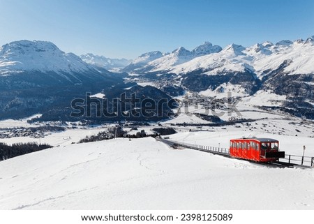 A funicular carriage climbs on the snowy slope toward Muottas Muragl, overlooking St Motritz, Celerina, Pontresina and lakes in Engadin Valley under alpine mountains, in Samedan, Grisons, Switzerland Royalty-Free Stock Photo #2398125089