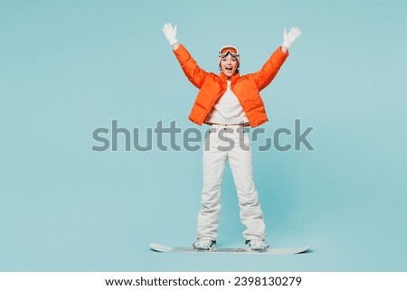 Full body young excited woman wear warm padded windbreaker jacket hat ski goggles mask snowboarding raise up hand travel rest spend weekend winter season in mountains isolated on plain blue background