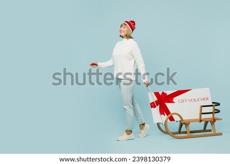 Full body merry elderly woman 50s years old wear sweater red hat posing carry gift certificate coupon voucher card for store on sled isolated on plain blue background. Happy New Year holiday concept
