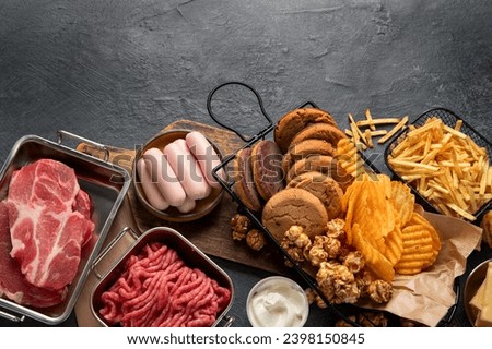 Saturated and trans. Unhealthy food. Sausage, potato, meat, cheese, popcorn, cookies, cream on dark background. Top view, copy space. Royalty-Free Stock Photo #2398150845