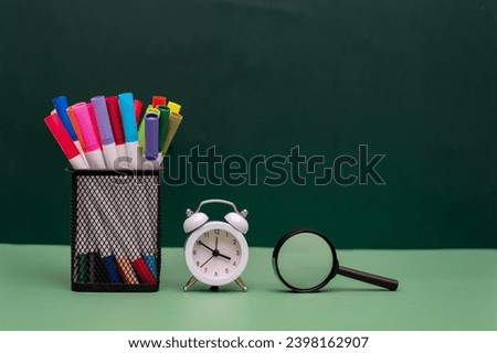 A bunch of colored pens in the square metal pen holder sitting next to white alarm clock and magnifying glass on green surface