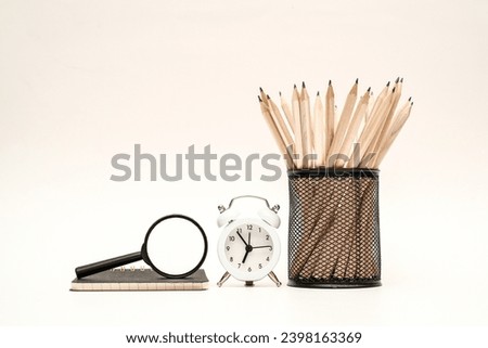 A bunch of pencils in the round metal pencil holder sitting next to black alarm clock and magnifying glass on white surface