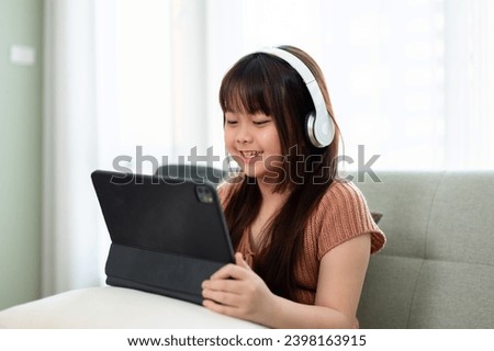 Young adorable and happy Asian girl in casual clothes wearing headphones and enjoying playing games or watching kid cartoons online on her digital tablet on a sofa in the living room.