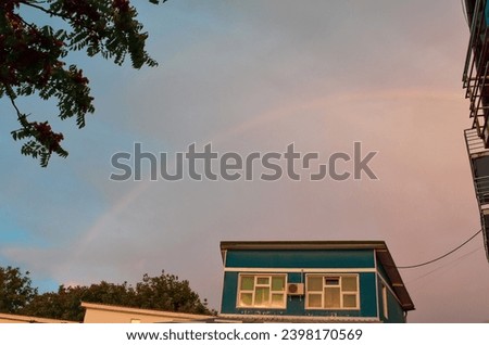 The sky after the rain with a rainbow in the background. Urban landscape with a blue building and trees behind the building
