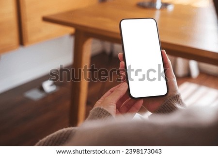 Mockup image of a woman holding mobile phone with blank desktop white screen at home
