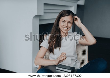 Relaxed young woman in casual clothes smiling wide sitting in chair looks at camera happily against fireplace using laptop for remote working. Cozy home, domestic education concept. Royalty-Free Stock Photo #2398177609