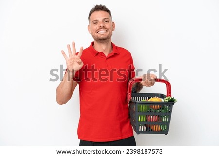 Young caucasian man holding a shopping basket full of food isolated on white background happy and counting four with fingers