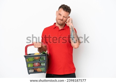 Young caucasian man holding a shopping basket full of food isolated on white background frustrated and covering ears Royalty-Free Stock Photo #2398187577
