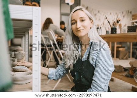 Woman entrepreneur wearing apron working pottery workshop and ceramic shop. Female craftsperson and owner of craft small business among handmade ceramic pottery objects in art studio workspace.
